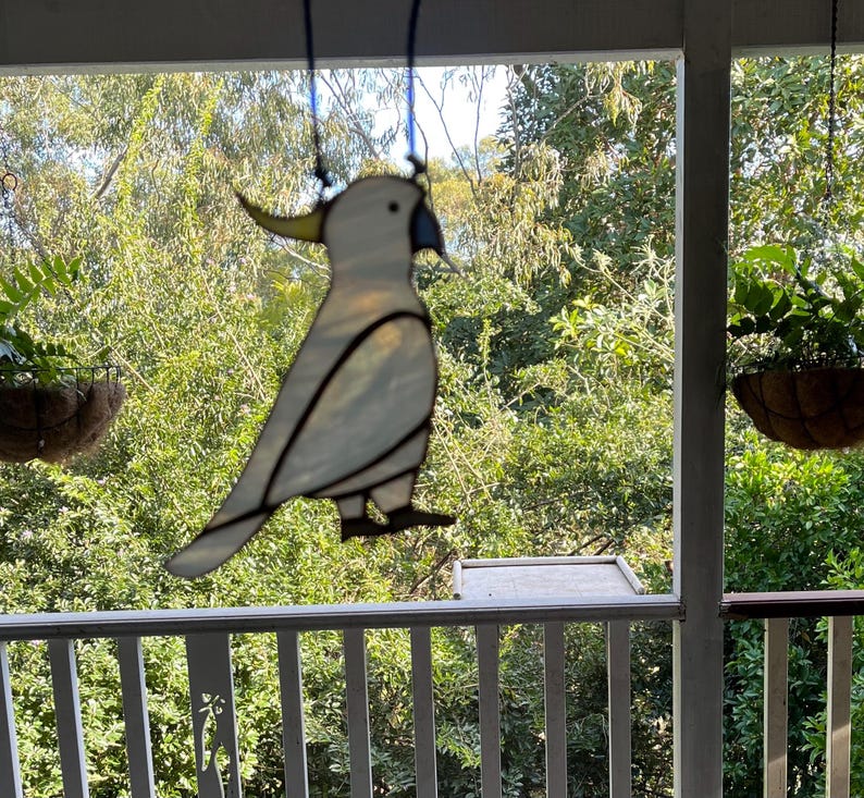May include: A white stained glass cockatoo bird hanging from a porch ceiling. The bird is suspended in front of a window with a view of green trees.