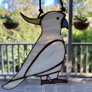 May include: A white stained glass cockatoo with a yellow beak and blue accents hanging from a black cord.