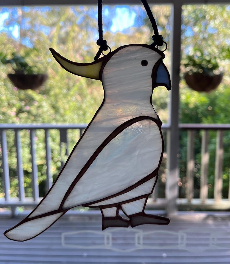May include: A white stained glass cockatoo with a yellow beak and blue accents hanging from a black cord.