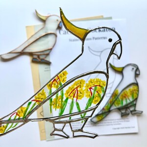 May include: A stained glass cockatoo bird with yellow, white, and red accents. The bird is decorated with yellow flowers and green leaves. The bird is on a white background with a printed pattern of a cockatoo.