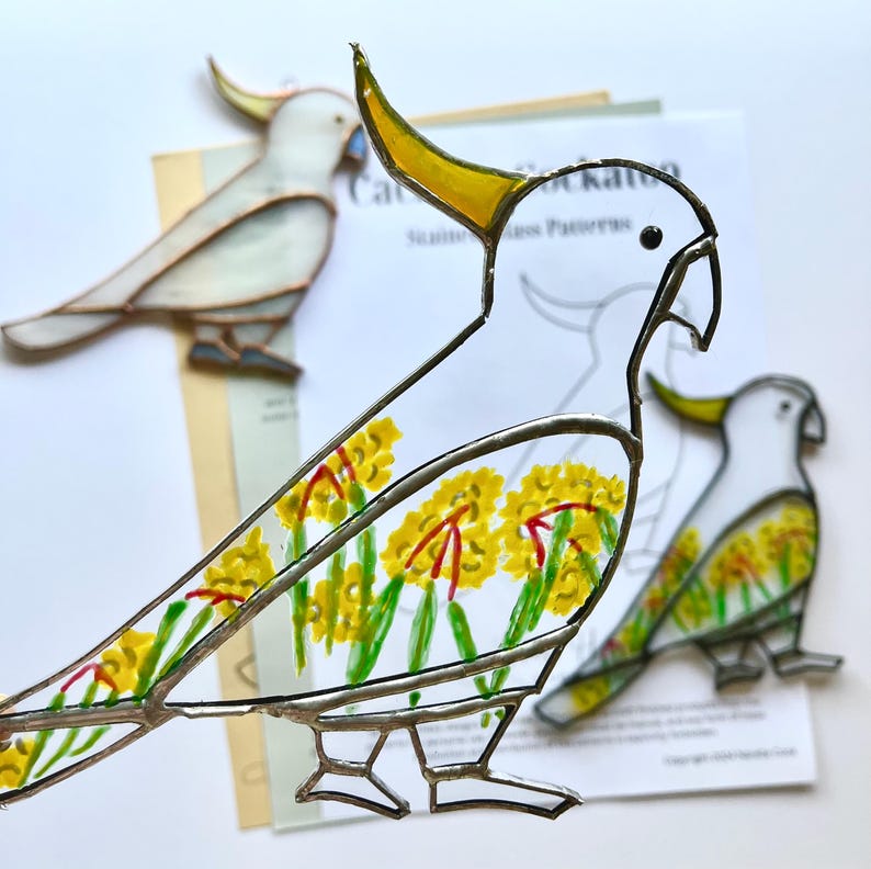 May include: A stained glass cockatoo bird with yellow, white, and red accents. The bird is decorated with yellow flowers and green leaves. The bird is on a white background with a printed pattern of a cockatoo.