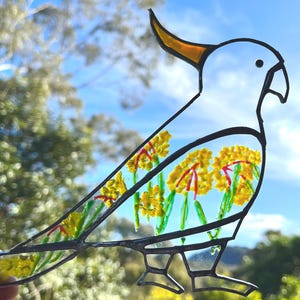 May include: A white stained glass cockatoo with a yellow crest, standing on two legs. The cockatoo is decorated with yellow and red flowers.