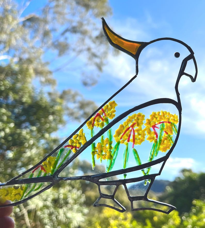 May include: A white stained glass cockatoo with a yellow crest, standing on two legs. The cockatoo is decorated with yellow and red flowers.