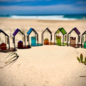 Könnte beinhalten: Eine Reihe von bunten Glashäusern auf einem Sandstrand. Die Häuser sind alle in verschiedenen Farben gehalten und haben unterschiedliche Designs. Sie sind in einer Reihe auf dem Sand angeordnet, mit dem Meer im Hintergrund.