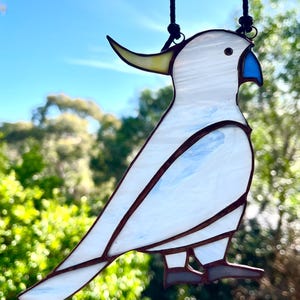 May include: A white stained glass cockatoo with a yellow crest and blue beak hanging from a black cord. The cockatoo is perched on a branch with its wings spread out.
