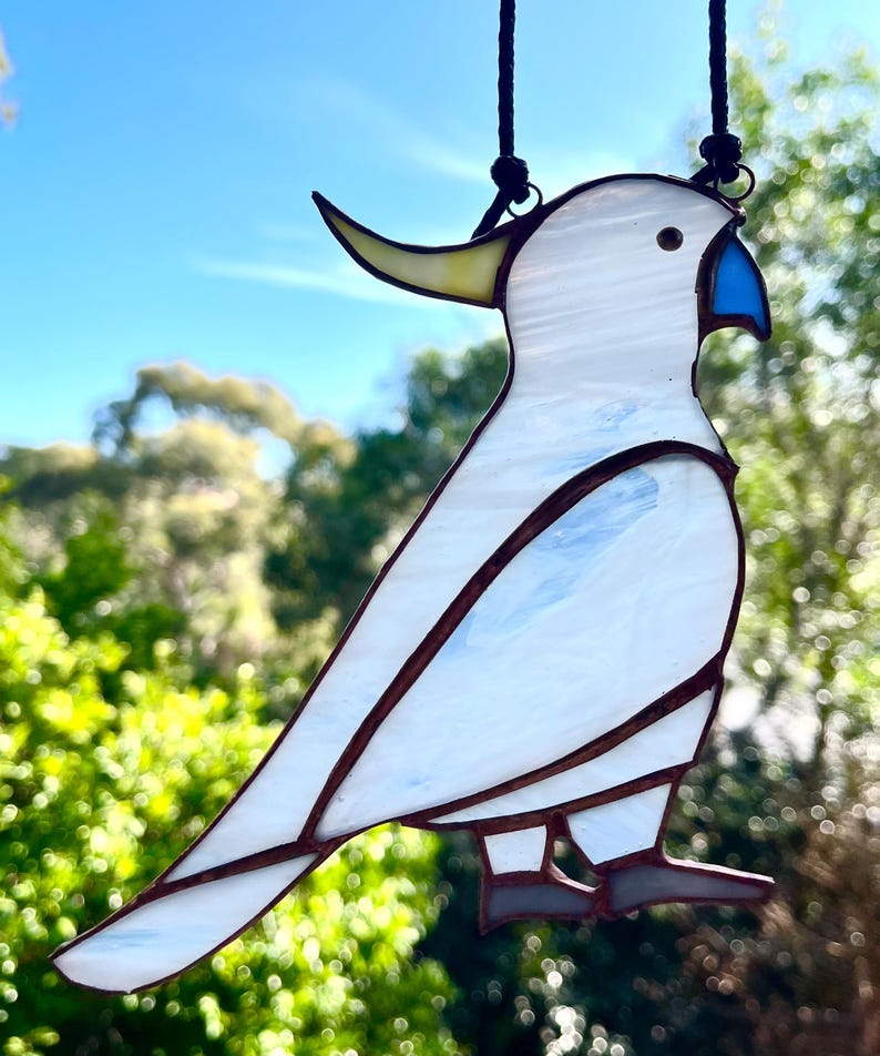 May include: A white stained glass cockatoo with a yellow crest and blue beak hanging from a black cord. The cockatoo is perched on a branch with its wings spread out.