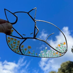 May include: A stained glass cat with a black outline, curled up with its head resting on its paws. The cat is surrounded by yellow and white flowers with green stems.