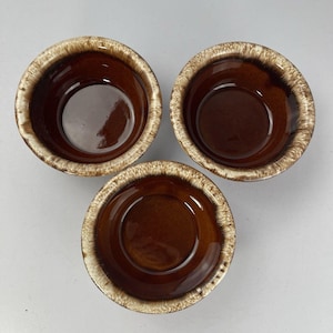 May include: Three brown ceramic bowls with a speckled, cream-colored rim. The bowls are round with a glossy interior and a matte exterior. The bowls are arranged in a triangular pattern against a neutral background. These bowls are suitable for serving food.