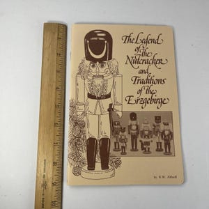 May include: A softcover book titled The Legend of the Nutcracker and Traditions of the Erzgebirge by K.W. Althoff, featuring a brown line drawing of a nutcracker. A wooden ruler shows the book is approximately 24 cm tall.