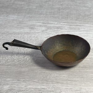 Puede incluir: Una pequeña sartén de cobre martillado con un mango largo y oscuro y un gancho en el extremo. La sartén tiene forma de cuenco redondeado y una pátina oscura y envejecida. El mango está sujeto a la sartén con remaches.