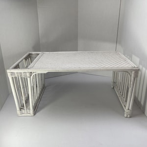 Puede incluir: Una bandeja de cama de mimbre blanco con una parte superior y laterales rectangulares tejidos. La bandeja tiene un borde elevado en un lado y un borde inferior en el otro. Las patas están en ángulo.