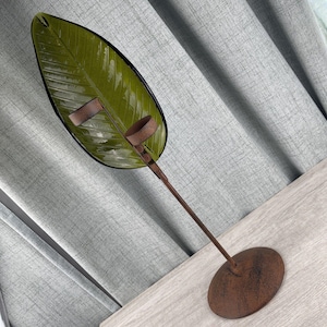Peut inclure: Un bougeoir décoratif composé d'une grande feuille en verre vert olive brillant fixée sur une fine tige en métal rouillé brun foncé. La tige repose sur une base circulaire en métal texturé brun foncé, formant une structure haute et droite.