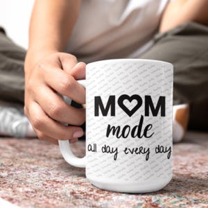 Peut inclure: Une tasse en céramique blanche avec du texte noir qui dit "MOM mode all day every day" avec un symbole de cœur dans le mot MOM.