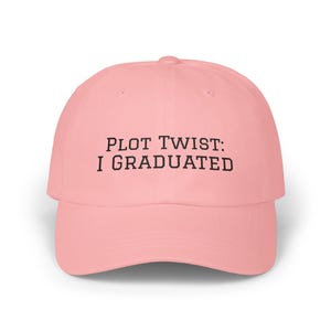 May include: A pale pink baseball cap with a curved peak. The cap displays the text "PLOT TWIST: I GRADUATED" in black, centrally positioned. The hat is made of a soft material and has a traditional six-panel design.