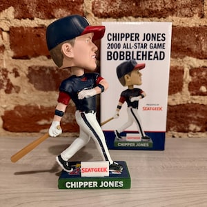 May include: A bobblehead of baseball player Chipper Jones in a blue and red Atlanta Braves uniform, holding a baseball bat. The bobblehead is on a green base with the words "SeatGeek" and "Chipper Jones" printed on it. The bobblehead is in front of a box with the words "Chipper Jones 2000 All-Star Game Bobblehead" printed on it.