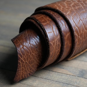 Op de afbeelding: Opgerold bruin leermateriaal met een getextureerd oppervlak. Het leer heeft een warme, aardse tint en de randen zijn netjes afgewerkt. Het materiaal lijkt flexibel en geschikt voor diverse knutselprojecten.