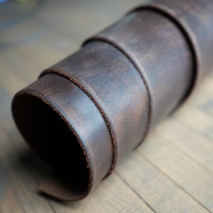May include: Rolled brown leather material, showing the texture and layers of the material. The leather has a distressed look, with visible grain and variations in color. The edges are neatly finished, suggesting a high-quality product.