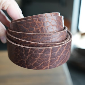 May include: A rolled-up, brown leather belt with a textured surface. The belt is a rich, dark brown color and appears to be made of high-quality leather. The edges of the belt are smooth and the leather has a natural grain.