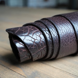 Puede incluir: Cuero marrón oscuro enrollado con una apariencia agrietada y texturizada. El cuero está enrollado firmemente, revelando las capas internas. Los bordes son lisos y oscuros, contrastando con el fondo de vetas de madera más claro. El cuero parece ser de alta calidad.