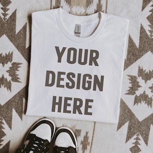 May include: White t-shirt with the text "YOUR DESIGN HERE" printed on the front. The t-shirt is lying flat on a patterned rug.