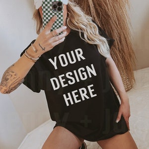May include: A black oversized t-shirt with the text "YOUR DESIGN HERE" printed in white. The shirt is being worn by a person with a white baseball cap and a checkered phone case.