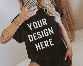May include: A black oversized t-shirt with the text "YOUR DESIGN HERE" printed in white. The shirt is being worn by a person with a white baseball cap and a checkered phone case.