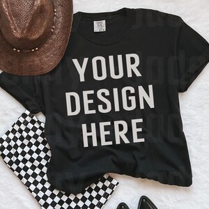 May include: Black t-shirt with the text "YOUR DESIGN HERE" printed in white. The t-shirt is laying flat on a white surface with a brown hat, black and white checkered pants, and black cowboy boots.