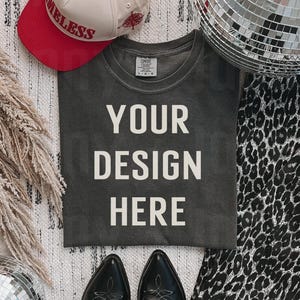 May include: A dark gray t-shirt with the text "YOUR DESIGN HERE" printed in white. The t-shirt is laying flat on a white and gray patterned surface. There is a red and white baseball cap, two black cowboy boots, and two disco balls in the image.