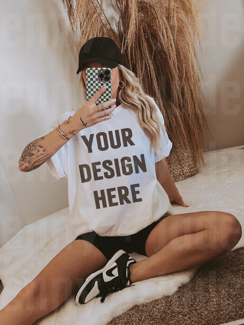 May include: A white t-shirt with the text "YOUR DESIGN HERE" printed on the front. The t-shirt is being worn by a person sitting on a white fluffy rug. The person is wearing a black baseball cap and black shorts.