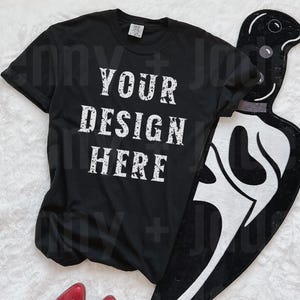 May include: Black t-shirt with the text "YOUR DESIGN HERE" in white lettering. The shirt is lying on a white surface with a black and white graphic design next to it.