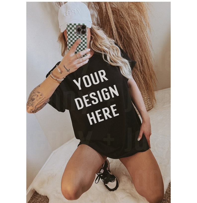 May include: A black oversized t-shirt with the text "YOUR DESIGN HERE" printed in white. The shirt is being worn by a person with a white baseball cap and a checkered phone case.