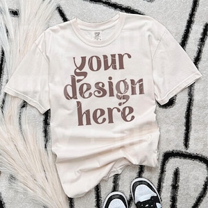 May include: A beige t-shirt with the text "your design here" printed in brown on the front. The shirt is laid flat on a white and black patterned surface.