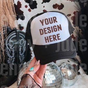 Puede incluir: Gorra de camionero negra con un panel frontal blanco. El panel presenta el texto "YOUR DESIGN HERE" en marrón. La gorra se sostiene frente a una bola de discoteca y una bota de vaquero negra.