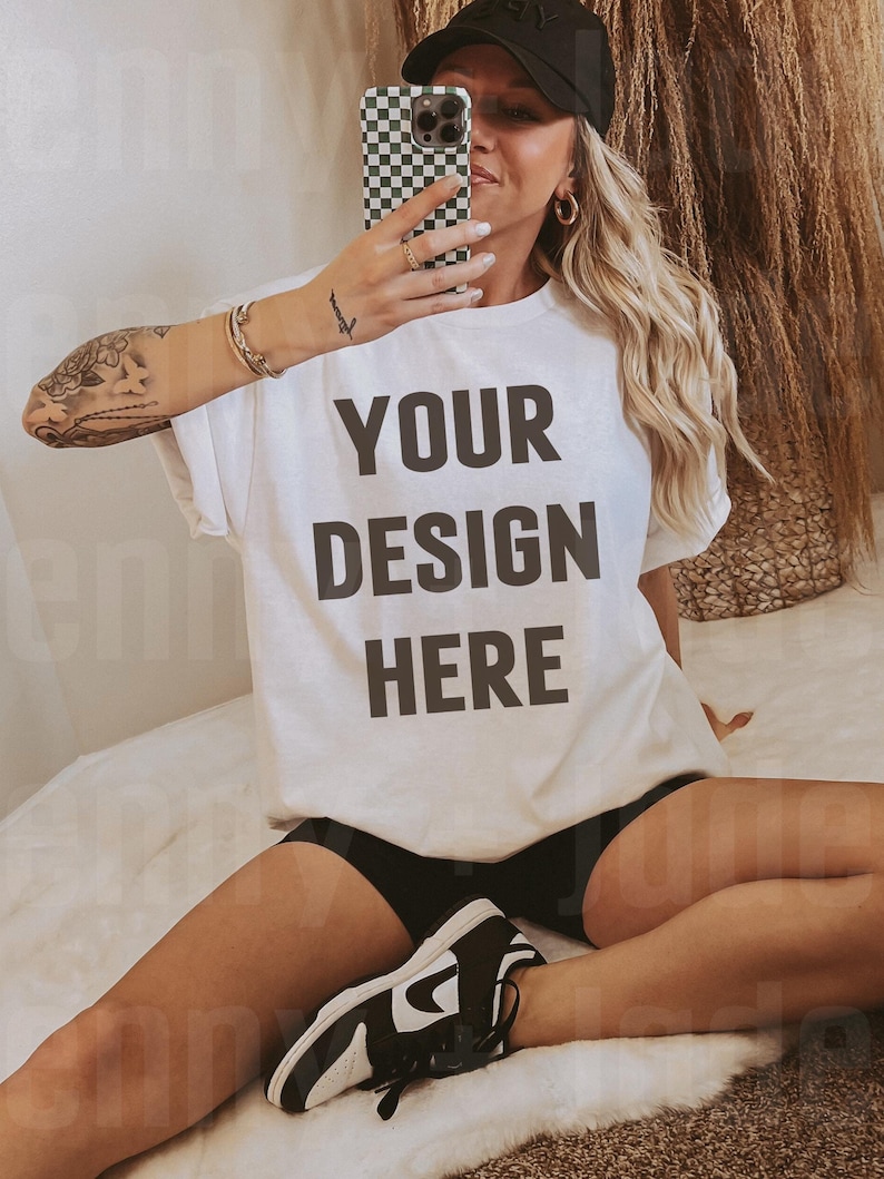 May include: A white t-shirt with the text "YOUR DESIGN HERE" printed in black. The shirt is being worn by a person sitting on a white fuzzy surface. The person is wearing black shorts and black and white sneakers.