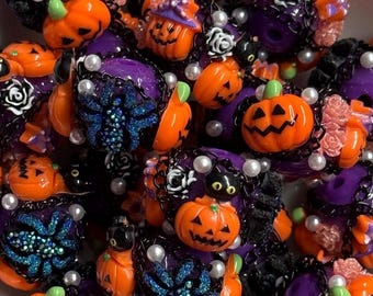 Può includere: Una collezione di perline a tema Halloween nei toni arancione, viola e nero. Le perline presentano jack-o'-lantern, ragni, gatti neri e accenti floreali. Alcune perline sono decorate con perle finte e dettagli a catena.