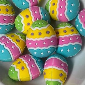 May include: A collection of colorful Easter egg decorations. Each egg features a vibrant design with sections of yellow, pink, green, and blue, accented with white wavy lines and small flower details. The eggs are arranged closely together.