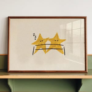 May include: A framed print featuring two yellow star characters with closed eyes and black outlines, resting on a black line. The left star has three black Zs above it. The print is housed in a rectangular wooden frame with a light-coloured mat.