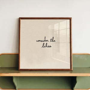 Puede incluir: Una impresión cuadrada enmarcada con un borde de madera oscura que presenta el texto cursivo negro "consider the lilies" sobre un fondo beige claro, apoyada en un estante de pared verde oliva con bordes redondeados y acabado en madera clara.