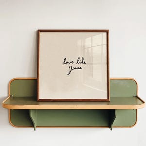 Puede incluir: Un estante de pared cuadrado con un panel trasero verde claro y un marco de madera clara, que sostiene una impresión cuadrada enmarcada. La impresión presenta un fondo beige con el texto cursivo negro "love like Jesus" centrado en el marco.