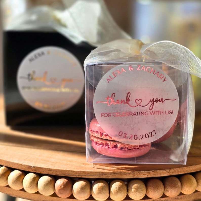 May include: A clear square favour box containing pink macarons, topped with a sheer white ribbon. A circular sticker on the front features rose gold text reading Alexa & Zachary, thank you for celebrating with us! and the date 03.20.2027.