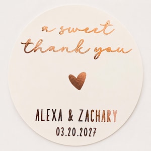 May include: A circular off-white sticker featuring copper-coloured foil text that reads a sweet thank you, a small copper heart, and the names ALEXA & ZACHARY followed by the date 03.20.2027 printed in a dark brown sans-serif font.
