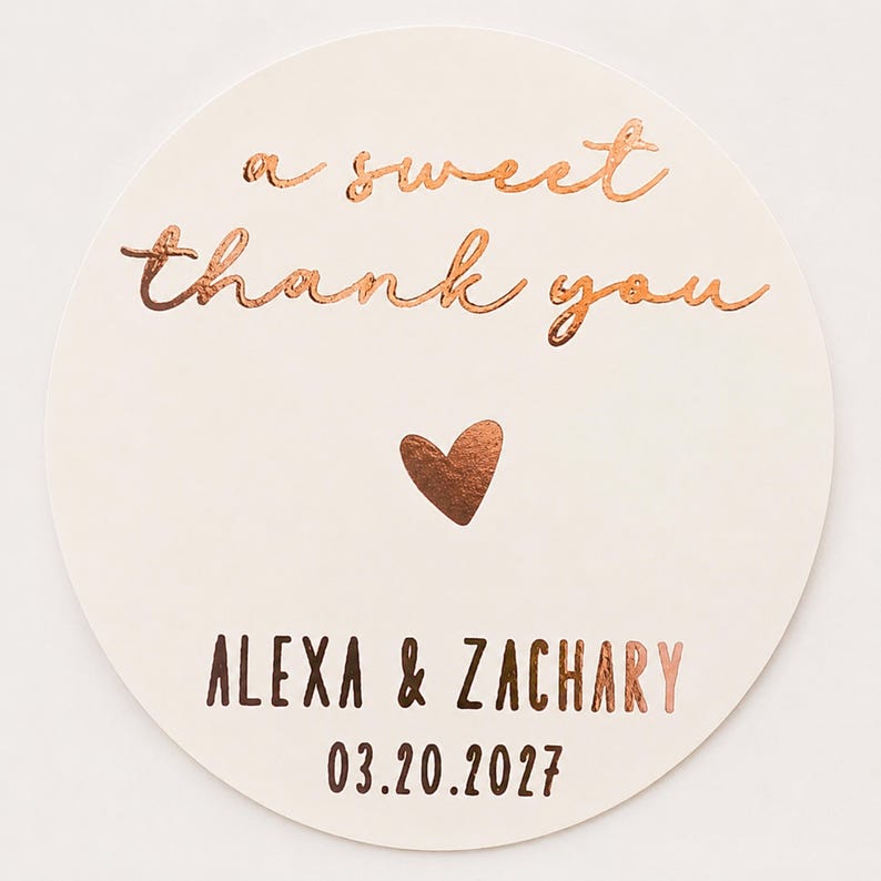 May include: A circular off-white sticker featuring copper-coloured foil text that reads a sweet thank you, a small copper heart, and the names ALEXA & ZACHARY followed by the date 03.20.2027 printed in a dark brown sans-serif font.