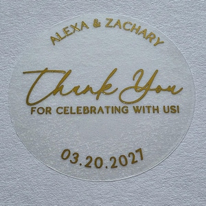 May include: A circular, translucent sticker featuring gold foil text. The top reads ALEXA & ZACHARY, the centre says Thank You, the bottom text reads FOR CELEBRATING WITH US!, and the date 03.20.2027 is printed at the very bottom in gold.
