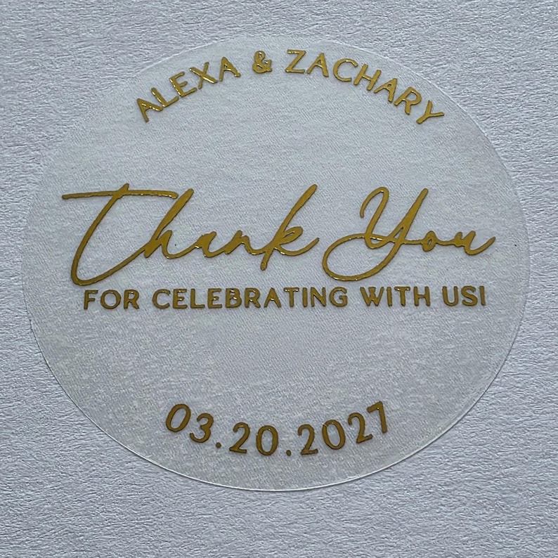 May include: A circular, translucent sticker featuring gold foil text. The top reads ALEXA & ZACHARY, the centre says Thank You, the bottom text reads FOR CELEBRATING WITH US!, and the date 03.20.2027 is printed at the very bottom in gold.