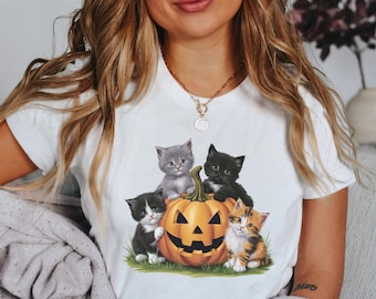 May include: A white t-shirt featuring a graphic of three kittens perched on a jack-o'-lantern. The kittens are grey, black, and orange and white.