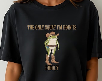 Op de afbeelding: Zwart oversized T-shirt met een afbeelding van een kikker die een cowboyhoed en laarzen draagt. De tekst "The only squat I'm doin' is diddly" staat op het shirt gedrukt.