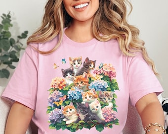 May include: Pink t-shirt featuring a design of a group of kittens surrounded by flowers. The kittens are various colours and have different expressions.