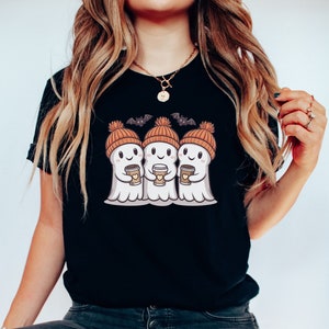 K&ouml;nnte beinhalten: Schwarzes T-Shirt mit einem Motiv von drei Geistern, die orangefarbene Strickm&uuml;tzen tragen und Kaffeetassen halten. Die Geister haben l&auml;chelnde Gesichter und sind von Flederm&auml;usen umgeben.