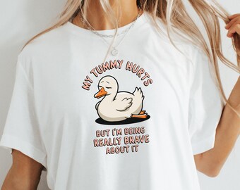 May include: White t-shirt featuring a cartoon duck with the text "MY TUMMY HURTS" above the duck and "BUT I'M BEING REALLY BRAVE ABOUT IT" below. The duck is white with an orange beak and feet.