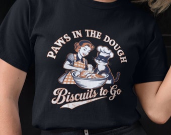 Può includere: Una maglietta nera con un'illustrazione in stile vintage di una donna e un gatto che fanno i biscotti. Il testo dice "Paws in the Dough Biscuits to Go".